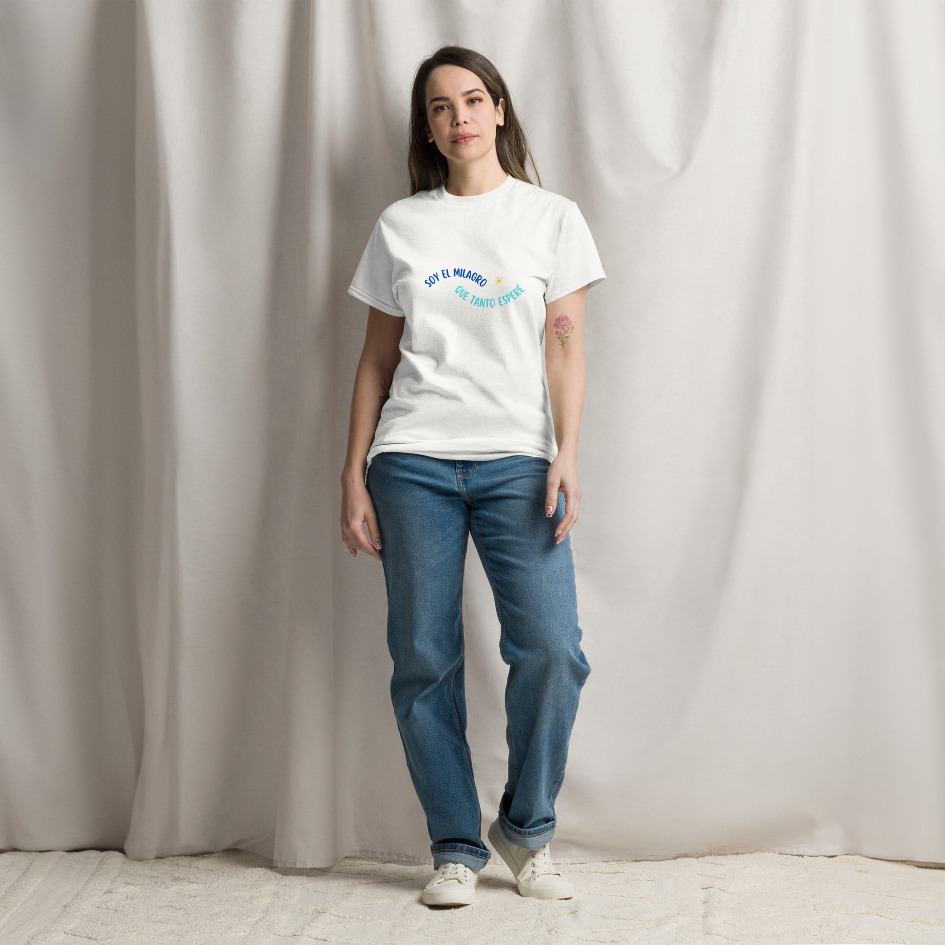 Mujer con camiseta blanca “Soy el milagro que tanto esperé” posando frente a cortina luminosa